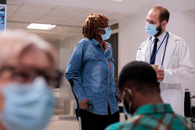 Afroamerikanischer Patient mit Krücken in der Krankenhauslobby, der mit einem männlichen Arzt über Gesundheitsversorgung und Genesung spricht. Frau mit chronischer Behinderung und Beeinträchtigung, die einen Kontrollbesuch macht.