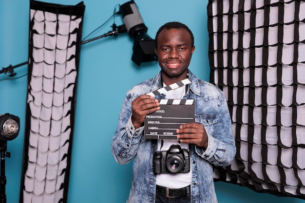 Afroamerikanischer Produzent mit Filmklappe im Studio mit professioneller Produktionsausrüstung. Lächelnder Fotograf mit Klöppel auf blauem Hintergrund, während er Softboxen und Scheinwerfer hat.