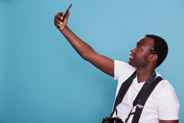 Afroamerikanischer professioneller Fotograf, der Selfie-Foto mit Smartphone-Gerät macht, während er auf blauem Hintergrund steht. Selbstbewusste Person mit DSLR-Kamera, während sie sich mit dem Telefon fotografiert.