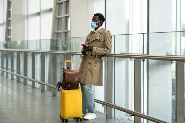 Afroamerikanischer Reisender Mann mit gelbem Koffer steht im Flughafenterminal, trägt eine schützende Gesichtsmaske, um sich vor dem Grippevirus, der Pandemie Covid-19, zu schützen und wartet auf Flug und Boarding.