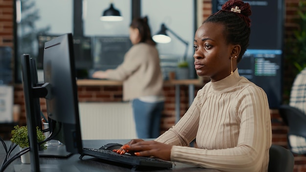 Afroamerikanischer Softwareingenieur, der konzentriert auf den Computerbildschirm blickt, während er in das Startup-Büro tippt. Datenbankprogrammierer, der Code mit Algorithmen schreibt, die auf Bildschirmen im Hintergrund kompiliert werden.