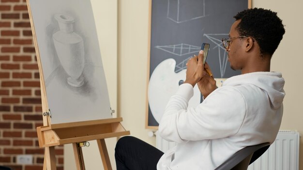 Afroamerikanischer Student, der mit dem Smartphone ein Bild vom Zeichnen in der Kunstunterrichtsschule macht und stolz auf die Skizzenpraxis ist. Erlernen neuer künstlerischer Fähigkeiten und Fotografieren von Kunstwerken.