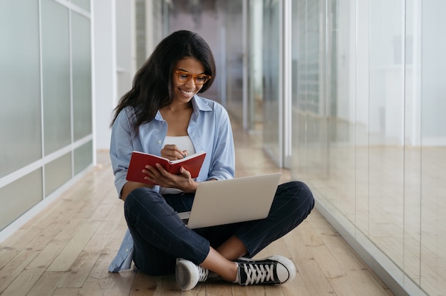 Afroamerikanischer Student, der mit einem Laptop studiert und sich Notizen macht. Glücklicher Freiberufler, der von zu Hause aus arbeitet