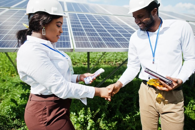 Afroamerikanischer Techniker überprüft die Wartung der Sonnenkollektoren. Gruppe von drei schwarzen Ingenieuren, die sich an der Solarstation treffen und einen Deal mit Geld machen.