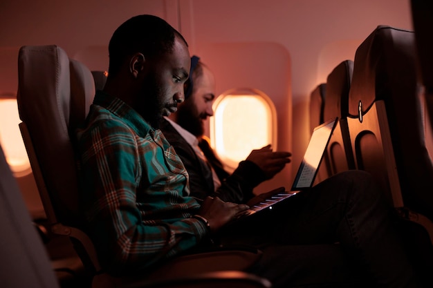 Afroamerikanischer Unternehmer, der während des Sonnenuntergangs am Laptop arbeitet und mit dem Flugzeug auf einem kommerziellen Flug mit internationalem Flugdienst fliegt. Männlicher Tourist, der auf Arbeitsreise oder Urlaub reist.