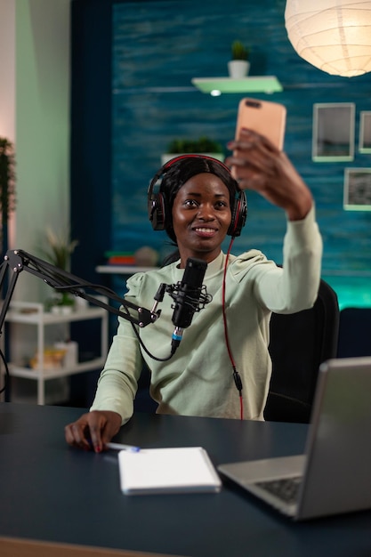 Afroamerikanischer Vlogger lächelt, während er während des Live-Streams ein Selfie mit einem modernen Smartphone macht. Ersteller von sozialen Medien, der ein Video-Tutorial mit Vlogging-Produktionsgeräten aufzeichnet. On-Air-Gespräch