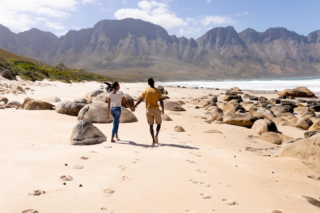 Afroamerikanisches Paar geht Händchen haltend an einem Strand am Meer spazieren. gesunder Lebensstil, Freizeit in der Natur.