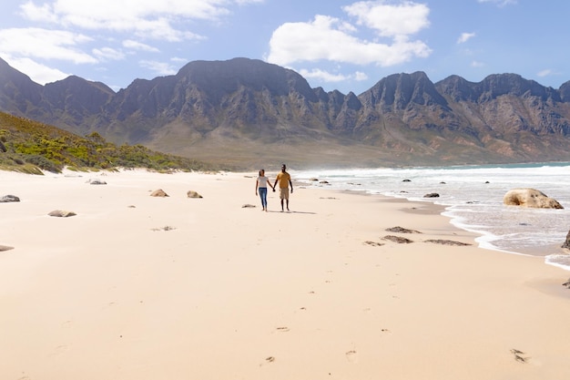 Afroamerikanisches Paar geht Händchen haltend an einem Strand am Meer spazieren. gesunder Lebensstil, Freizeit in der Natur.