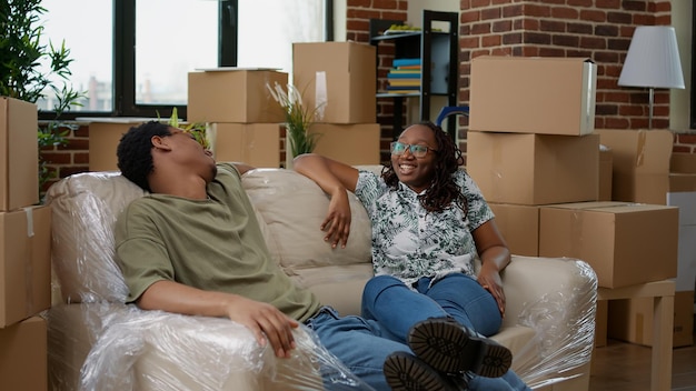 Afroamerikanisches Paar sitzt auf dem Sofa, um eine Pause vom Umzug in einen neuen Haushalt zu machen, der auf Leihbasis gekauft wurde. Auspacken von Möbeln in vermieteter Immobilienwohnung für Umzug, Neuanfang.