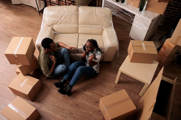 Afroamerikanisches Paar träumt von Wohnungsdekor, zieht in gemietete Haushaltswohnung und sitzt zusammen. Umzug ins Familienhaus, um neue Anfänge zu beginnen, Lebensereignis. Draufsicht auf.