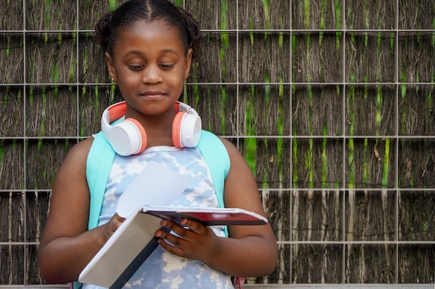 Afroamerikanisches Schulmädchen mit Rucksackkopfhörern und Notizbüchern am Schuleingang