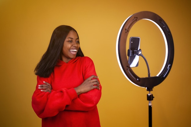 Afroamerikanisches Vlogger-Mädchen in einem roten Hoodie mit einer beleuchteten Ringlampe und verschränkten Armen des Telefons