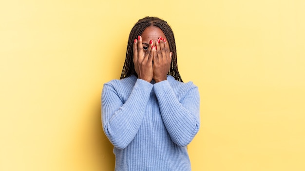 Afroschwarze hübsche Frau, die das Gesicht mit den Händen bedeckt, mit überraschtem Ausdruck zwischen den Fingern späht und zur Seite schaut