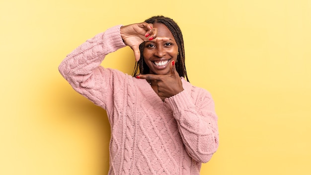 Afroschwarze hübsche Frau, die sich glücklich, freundlich und positiv fühlt, lächelt und ein Porträt oder einen Fotorahmen mit den Händen macht