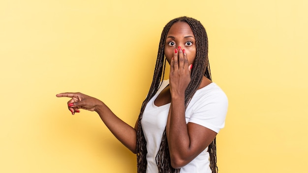 Afroschwarze hübsche Frau, die sich glücklich, schockiert und überrascht fühlt, den Mund mit der Hand bedeckt und auf den seitlichen Kopierraum zeigt