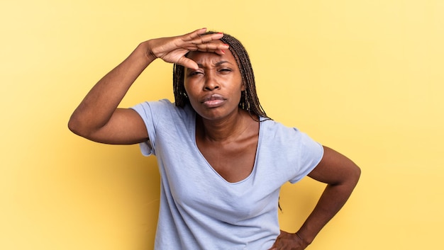 Afroschwarze hübsche Frau, die verwirrt und erstaunt aussieht, mit der Hand über der Stirn, die weit weg schaut, zuschaut oder sucht