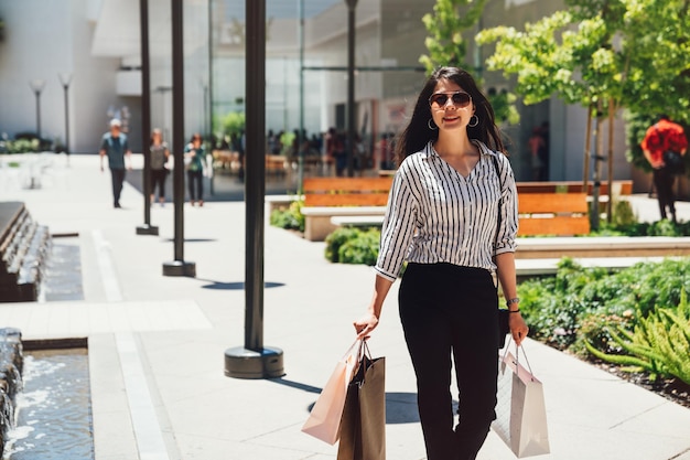 After-Day-Shop-Shop-Kauf am Boxing Day Sale. fröhliche junge asiatin, die einkaufstaschen trägt, während sie die straße im vordergrund außerhalb des stanford-einkaufszentrums entlang geht. Dame genießt Sonnenlicht.