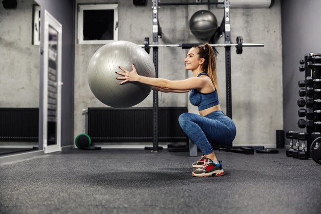 Agachamento com uma bola de fitness Uma mulher esbelta em roupas esportivas faz exercícios para queimar os músculos