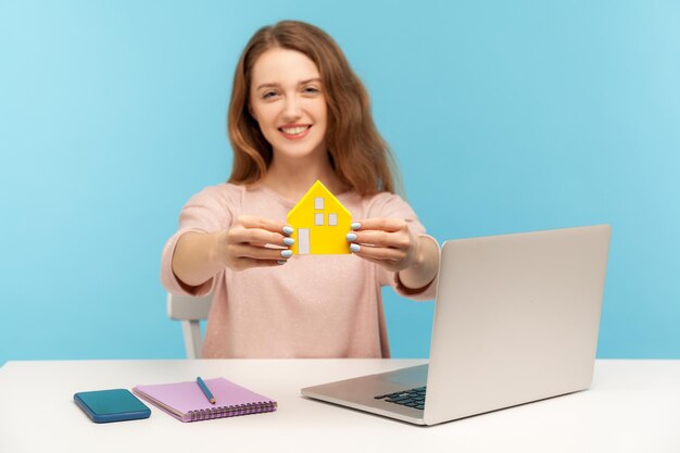 Agência imobiliária, compra de casa. Mulher alegre segurando a casa de papel e sorrindo para a câmera, sentada no local de trabalho com o laptop, trabalhando em home office. tiro de estúdio interno isolado em fundo azul