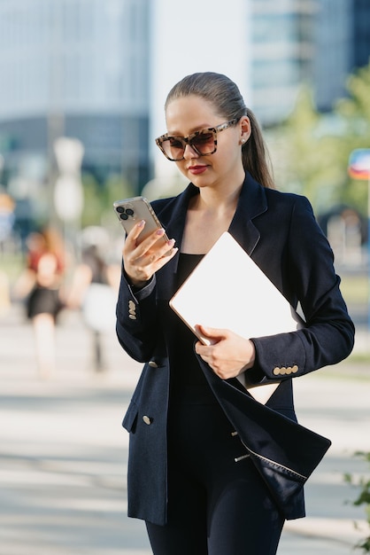 Una agente de bienes raíces con un blazer azul marino sostiene una computadora portátil entre los edificios de oficinas en el distrito financiero Una mujer de negocios está usando su teléfono inteligente en un grupo de edificios altos