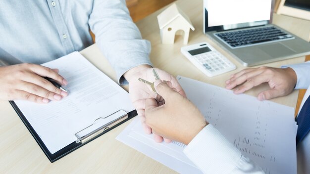 Agente de bienes raíces en traje sentado en un escritorio de oficina Entrega de llaves de casa con el cliente después de la firma del contrato