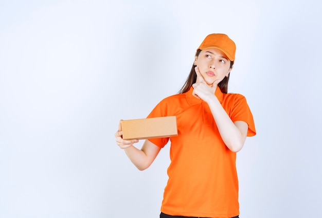 Agente de serviço feminino em dresscode cor laranja segurando uma caixa de papelão, parece pensativa e sonhando.