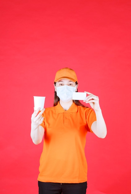 Agente de serviço feminino em uniforme de cor laranja e máscara, segurando um copo descartável e apresentando seu cartão de visita.