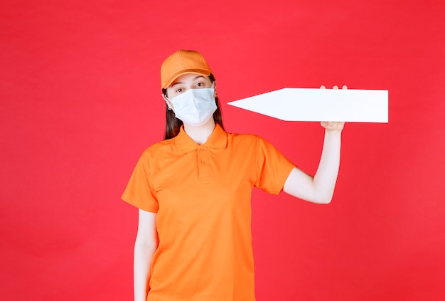 Agente de serviço feminino em uniforme de cor laranja e máscara segurando uma seta apontando para a esquerda.