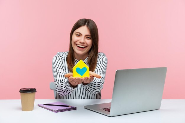 Agente imobiliário de mulher positiva sorridente segurando o modelo de casa de papel com coração nas mãos, sentado no local de trabalho com a construção de vida de segurança do laptop Tiro de estúdio interior isolado no fundo rosa