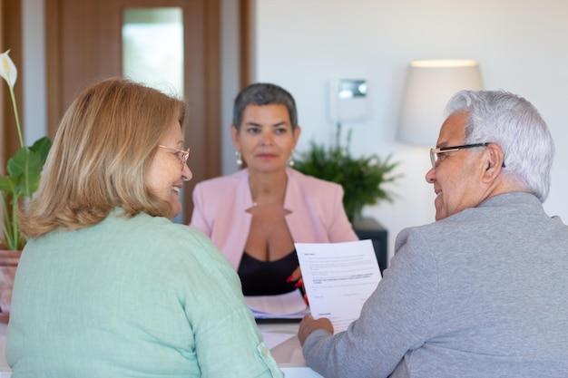 Agente inmobiliario enfocado que firma documentos con los clientes. Mujer de pelo corto con traje rosa sentada a la mesa con una pareja de ancianos, hablando, sonriendo. Bienes raíces, negocios, concepto de trabajo.