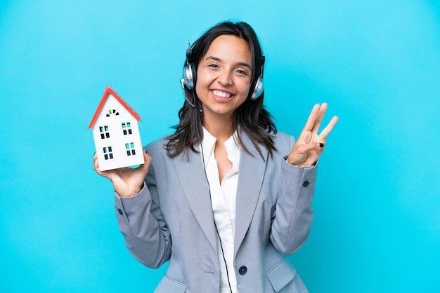 Agente inmobiliario hispano sosteniendo una casa de juguete aislada de fondo azul feliz y contando tres con los dedos