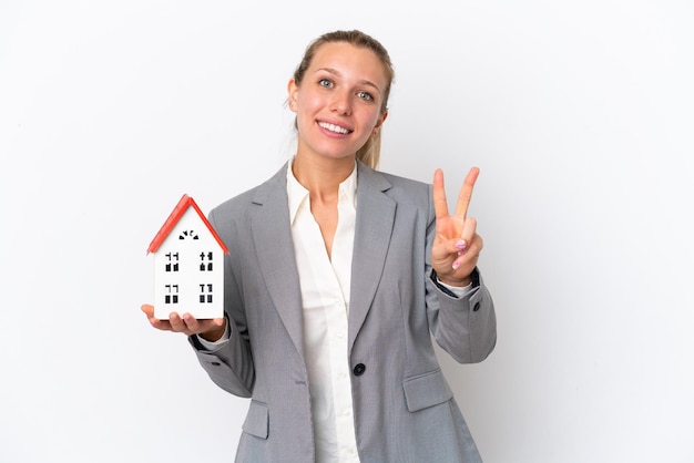 Agente inmobiliario mujer sosteniendo una casa de juguete aislada sobre fondo blanco sonriendo y mostrando el signo de la victoria