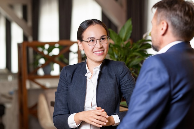 Agente muito jovem e alegre em trajes formais e óculos, olhando para o parceiro de negócios ou cliente durante a discussão de trabalho