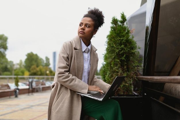 Agente de seguros de mujer afroamericana que trabaja en una computadora portátil en línea contra el fondo de la construcción de la ciudad