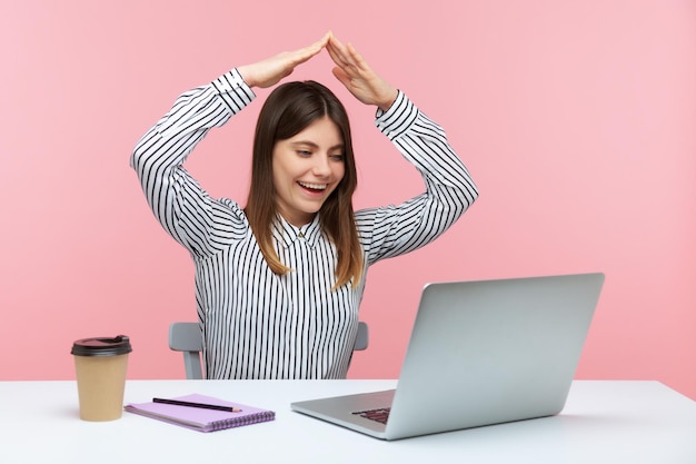 Una agente de seguros sonriente que muestra un gesto en el techo mirando la pantalla de un portátil hablando de una videollamada sentada en el lugar de trabajo en la oficina del hogar. Disparo de estudio interior aislado sobre fondo rosa