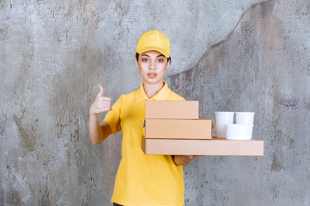 Foto agente de servicio femenino en uniforme amarillo sosteniendo un stock de cajas de cartón para llevar y vasos de plástico mientras muestra un signo de mano positivo