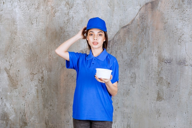 Foto agente de servicio femenino en uniforme azul sosteniendo un recipiente de plástico y parece confundida y pensativa o con una buena idea.