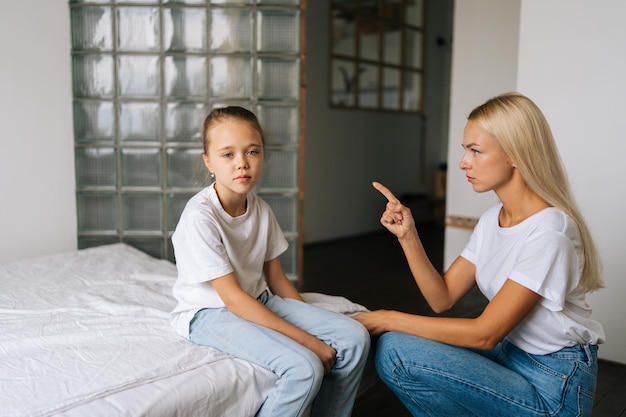 Aggressive junge Mutter, die mit erhobener Stimme schimpft und mit den Fingern auf die hartnäckige, schwierige kleine Tochter zeigt, die traurig in die Kamera schaut