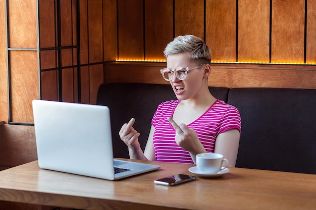 Aggressive unglückliche junge Freiberuflerin mit blonden kurzen Haaren in rosa T-Shirt und Brille sitzt im Café und hat schlechte Laune und zeigt Ficksingen mit dem Finger durch eine Webcam im Innenbereich