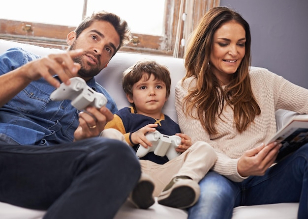 Agora temos que passar para a próxima fase Foto de um filho e seu pai jogando videogame juntos enquanto sua mãe lê uma revista