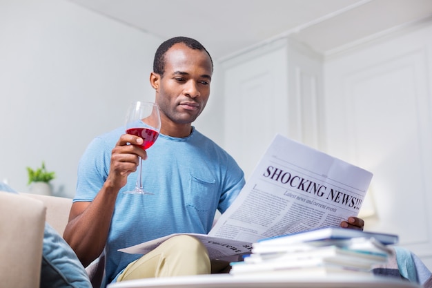 Agradable agradable guapo bebiendo vino y leyendo un periódico mientras descansa en casa