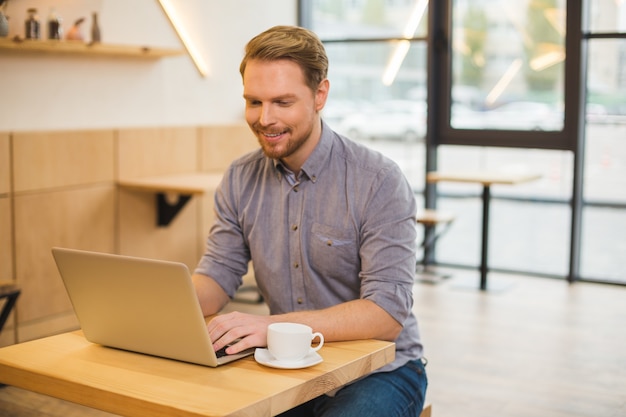 Agradable alegre hombre positivo sonriendo y mirando la pantalla del portátil mientras escribe