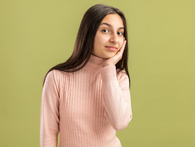 Agradable bastante adolescente mirando al frente manteniendo la mano en la cara aislada en la pared verde oliva con espacio de copia