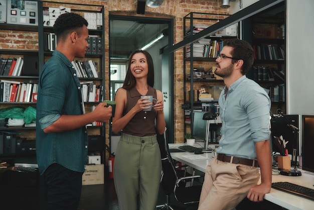 Agradable café jóvenes colegas sonrientes en ropa casual sosteniendo tazas y hablando de algo mientras