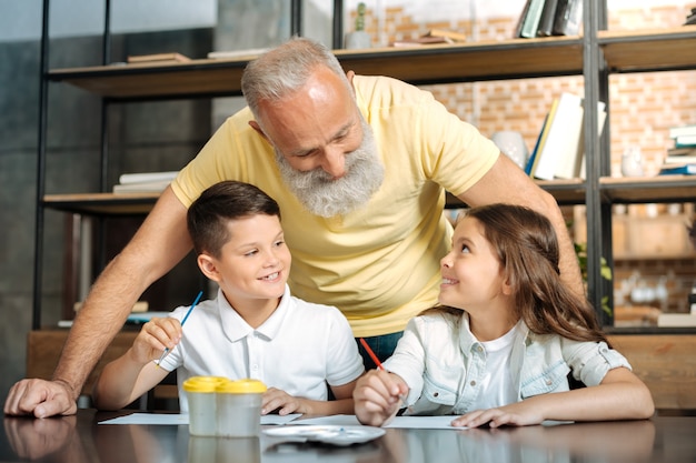 Agradable conversación. Dulce niño y niña preadolescentes sentados a la mesa, sosteniendo pinceles para pintar y hablando con su abuelo cariñoso