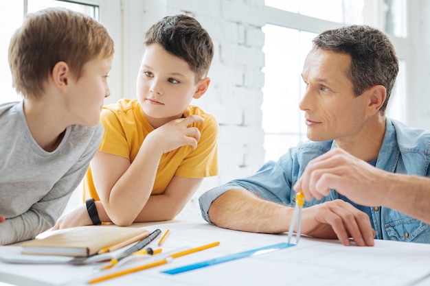 Agradable conversación. Encantadores niños preadolescentes sentados a la mesa en la oficina de su padre y hablando con él mientras él trabaja en un plano y usa una brújula