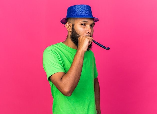 Agradable joven afroamericano con sombrero de fiesta soplando silbato de fiesta aislado en la pared rosa