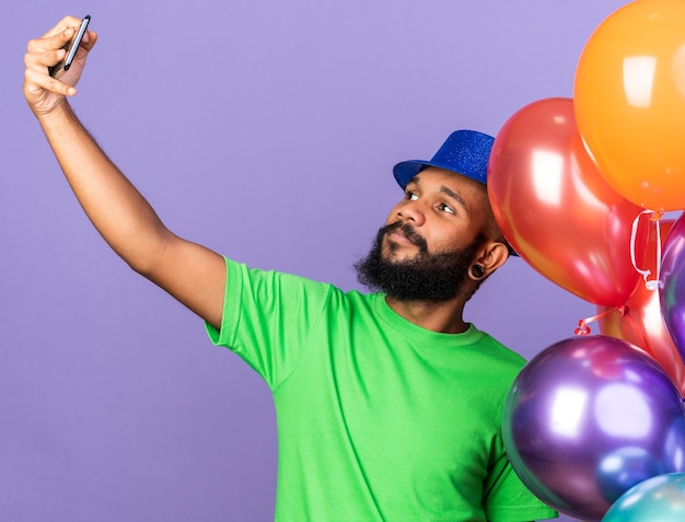 Agradable joven afroamericano con sombrero de fiesta sosteniendo globos tomar un selfie aislado en la pared azul