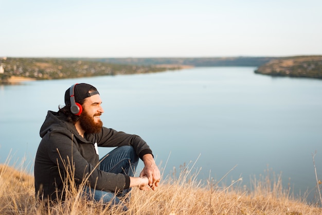 Agradable joven barbudo sentado en el campo escuchando música y sonriendo y mirando a otro lado