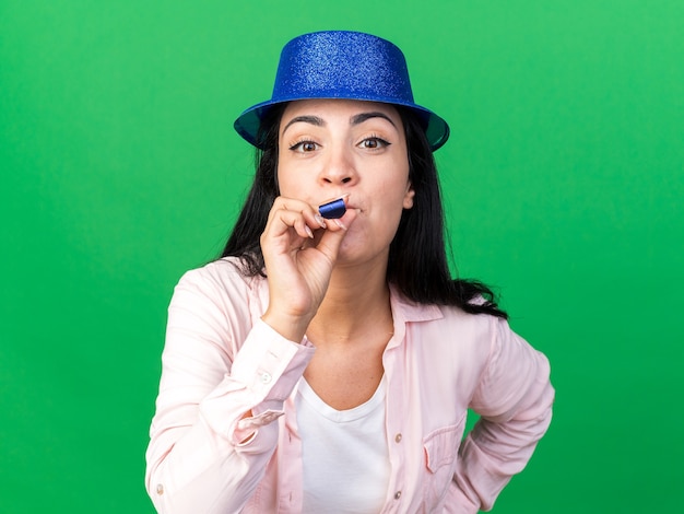 Agradable joven hermosa mujer vistiendo gorro de fiesta soplando silbato de fiesta poniendo la mano en la cadera